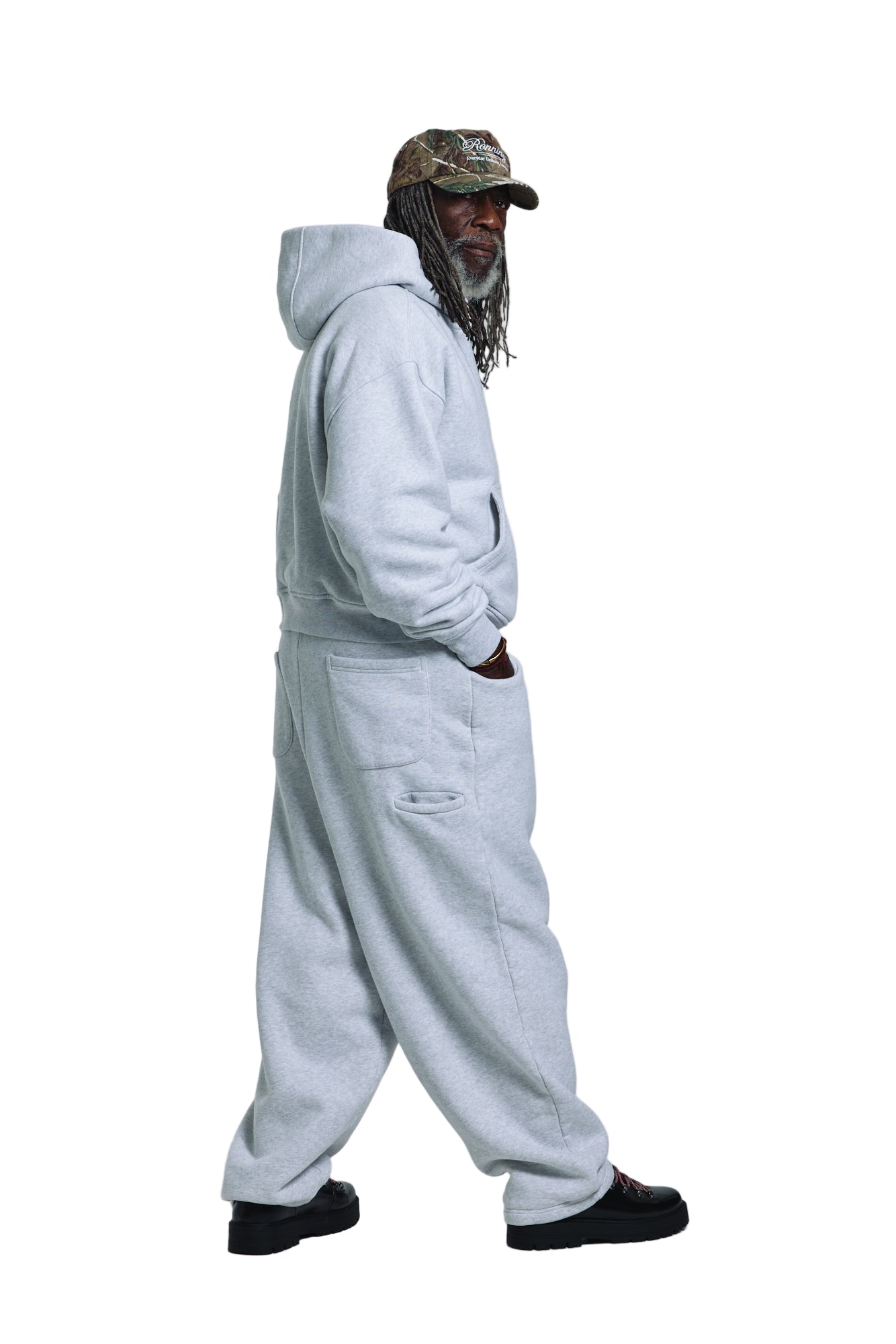 Person wearing a light gray hoodie and pants with a camouflage cap on a white background