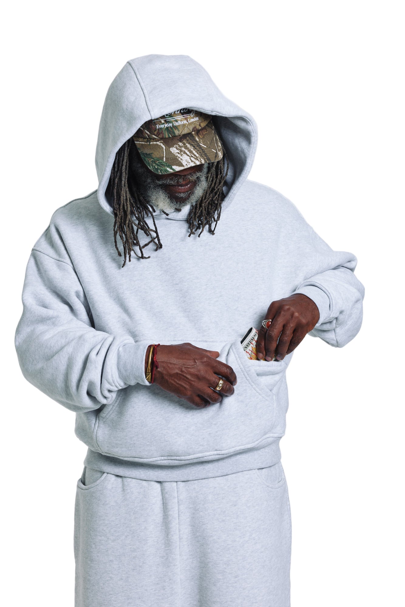 Person wearing a gray hoodie and pants with a camouflage cap, standing against a white background.